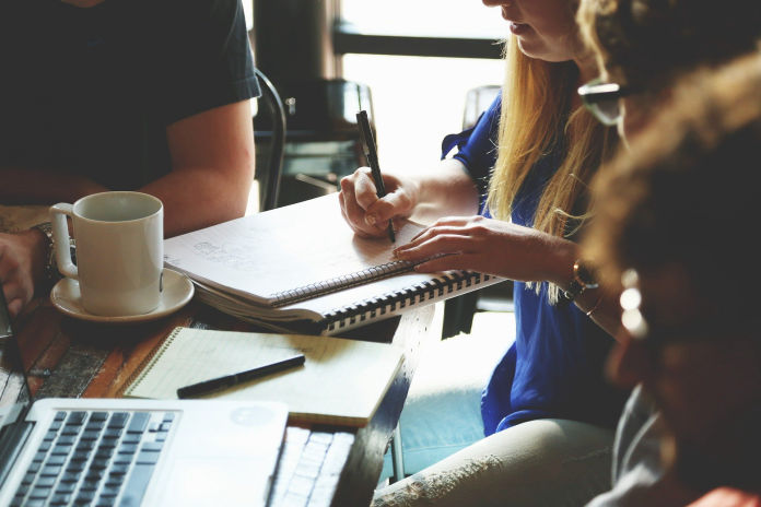 Arbeitsplatz mit Tastatur, Schreibheft, Kaffeetasse und 3 Personen, die dort sitzen